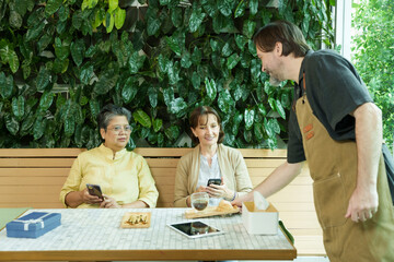 Asian senior woman and Caucasian woman sit at a cafe table with smartphones, smiling as a male waiter approaches, enjoying a joyful and relaxed retirement moment over tea and pastries.