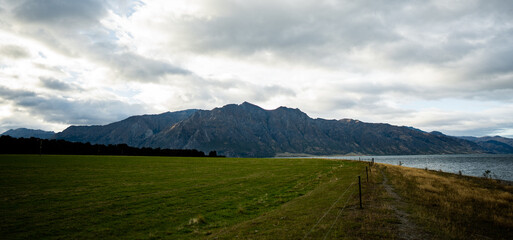 A vast green field leads to rugged mountains under a dramatic sky. Sunlight breaks through clouds, casting a peaceful glow over the landscape, evoking tranquility, grandeur, and natural harmony.