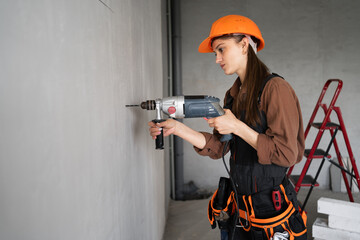 Female repair worker in helmet with tool belt drilling wall with electric drill during DIY home renovation