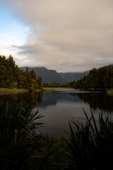 A serene lake reflects a misty mountain range, surrounded by lush greenery. Tall grasses frame the foreground, while clouds drift above, creating a peaceful and timeless natural scene.
