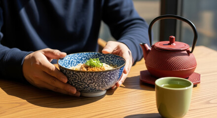 Person enjoying traditional japanese ramen with green tea and red teapot on sunlit wooden table