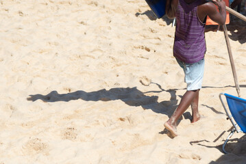 A worker at Porto da Barra beach is seen on the sand carrying material during strong sunlight. City of Salvador, Brazil.