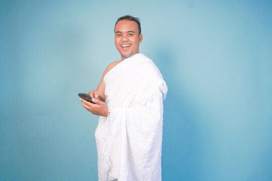 young Asian Muslim man wearing ihram doing video call pose while holding smartphone, isolated blue background. Hajj concept. islam