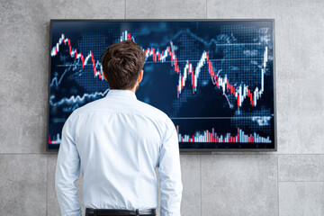 A man in a white shirt studies a large screen displaying fluctuating stock market charts and data.