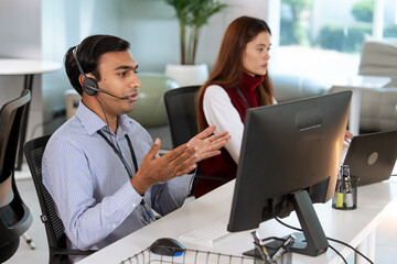 Customer support team working in modern office, with male representative wearing headset and gesturing while assisting customer, and female colleague focused on her laptop