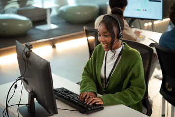 Cheerful customer support representative wearing headset and green sweater is working at computer in modern office environment, providing tech assistance