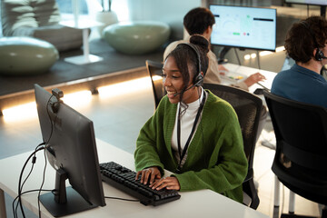 Cheerful customer support representative wearing headset is working on computer in modern office environment. Other team members are visible in background, also engaged in tech related tasks