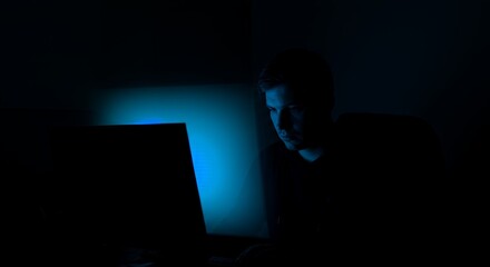 Late-night work session illuminated by a laptop's blue glow, a young man intensely focused on his task in the dark.