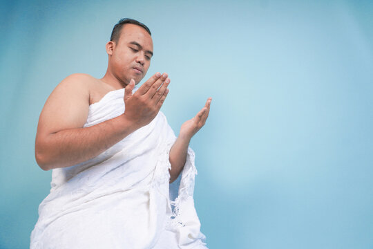 Young serious Asian Muslim man wearing ihram sitting and praying holding hands up with hand gesture isolated on blue background. Hajj concept. islam muslim concept