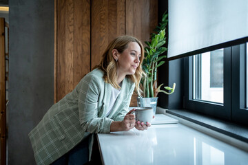 Businesswoman drinking coffee and looking out the window in modern office