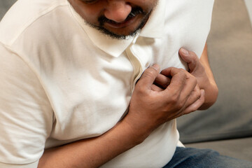 Asian man clutching chest in pain, showing signs of heart attack or serious health issue at home.