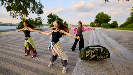 Sound in motion. Group of energetic young women practice street dance by a riverbank. Concept of urban fitness, community bonding and youth music education