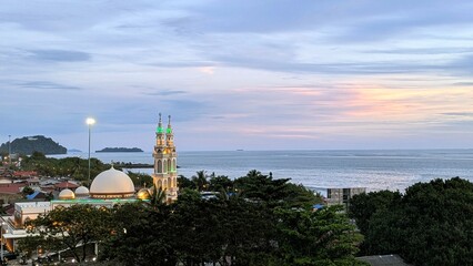 Fototapeta premium A high-resolution photograph of a beautifully lit mosque near the coastline during sunset. The mosque features illuminated domes and tall minarets, surrounded by palm trees and rooftops.