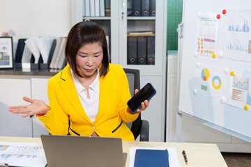 Confused businesswoman in yellow jacket holding phone and looking at laptop in office