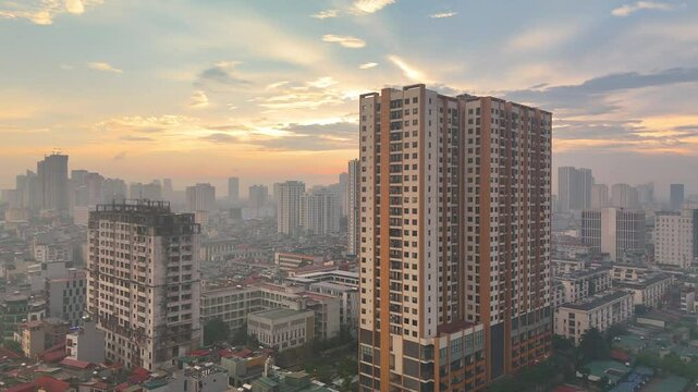An atmospheric aerial view of Hanoi, Vietnam, at sunset, bathed in the warm glow of the sun through a soft haze. The sprawling urban landscape stretches towards the horizon, with distant lakes or rive