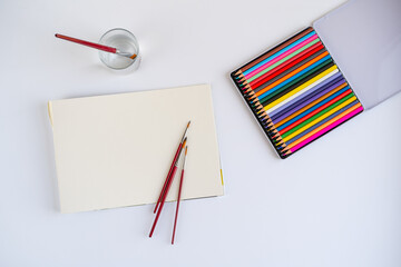 Creative workspace setup with color pencils, brushes, sketchbook, blank paper, and a water cup –...