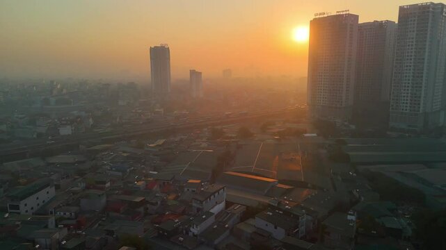 An atmospheric aerial view of Hanoi, Vietnam, at sunset, bathed in the warm glow of the sun through a soft haze. The sprawling urban landscape stretches towards the horizon, with distant lakes or rive