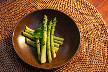 Asparagus spears arranged in a brown bowl, garnished with a green leaf, resting on a woven mat.