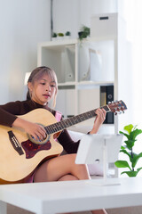 Young Asian woman playing acoustic guitar looking at tablet for online lesson at home studio in...
