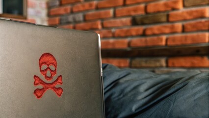A laptop with a red skull and crossbones embroidered on the cover sits on a black surface against a brick wall background.
