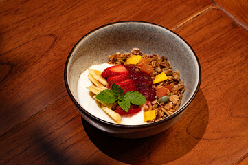 A bowl of yogurt topped with granola, sliced strawberries, bananas, jam, dried fruit, and a mint sprig sits on a wooden surface.