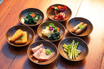 Seven small bowls filled with various breakfast foods, including ham, sausages, spinach, and potatoes, arranged on a wooden table.