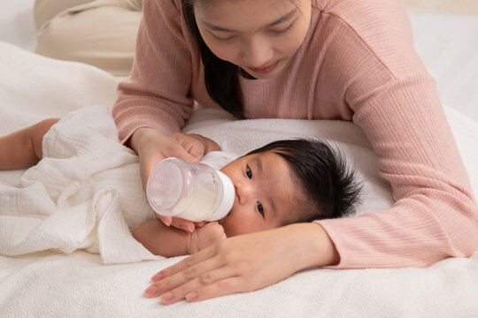 Mother, baby, newborn, family, love shown as little infant calmly drinks from bottle held by young Asian mom, reflecting softness, nurture, trust, peaceful care, and tenderness in relaxed setting