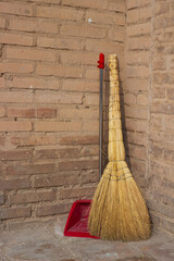 Close-up of a natural sorghum broom and red plastic dustpan in the corner by the brick wall. Concept background of hygiene and home cleaning.