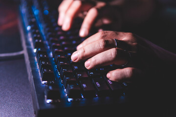 Close-up hands typing on a mechanical keyboard with LED backlight in a dark room. Shallow depth of...