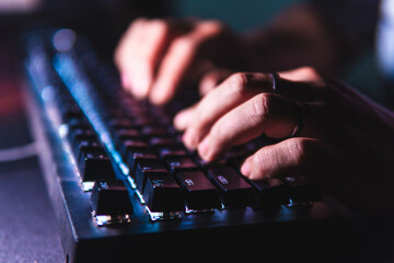 Close-up hands typing on a mechanical keyboard with LED backlight in a dark room. Shallow depth of...