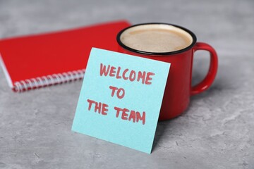 Sticky note with phrase Welcome To The Team, coffee and notebook on grey table, closeup
