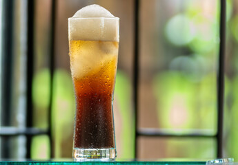 A glass of light beer, with foam, rests on a glass surface, with a softly blurred background.
