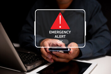 A person holds a smartphone displaying an emergency alert notification, with a laptop and tablet...