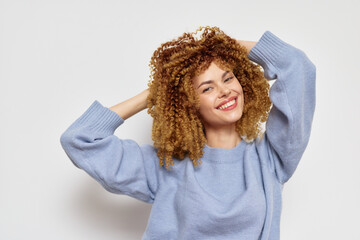 Smiling woman with curly hair wearing a light blue sweater, posing playfully against a neutral...