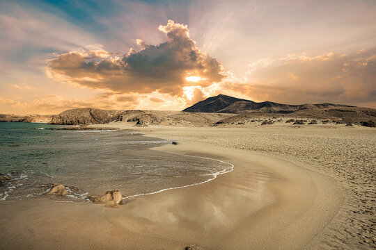 Sandstrand mit H&uuml;geln im Hintergrund bei untergehender Sonne, die teilweise von einer Wolke verdeckt wird.
