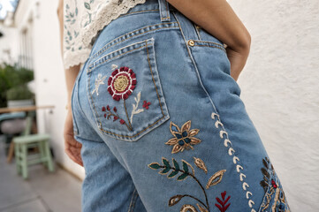 shows a close-up of a person wearing embroidered jeans, with intricate floral designs on the back pocket and down the side. The background features a white wall and some outdoor furniture.