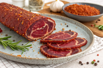 A plate of sliced meat, likely salami, is adorned with rosemary sprigs and seasoned with peppercorns and garlic. In the background, there is a bowl of red seasoning and a glass container of McCormick 