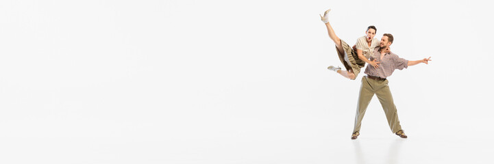 A couple performs an energetic swing dance move in vintage attire against a white background....