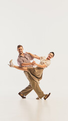 A couple executes an energetic dance move, dressed in vintage attire, against a plain backdrop....