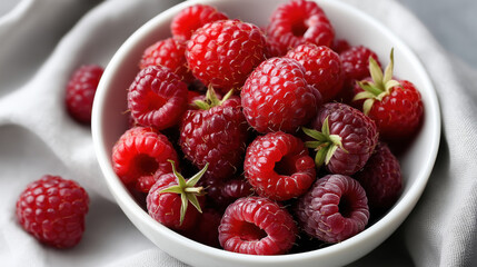 Fresh ripe raspberries in white bowl on soft fabric background. Bright red berries showcase natural summer harvest and healthy organic food.