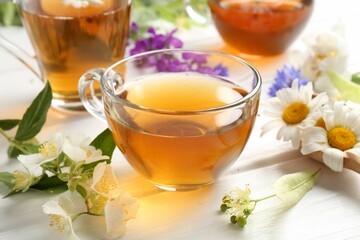 Aromatic herbal tea in glass cups and different flowers on white wooden table, closeup