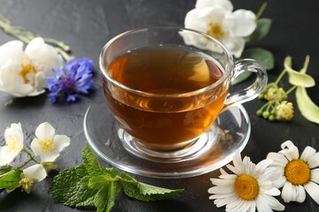 Aromatic herbal tea in glass cup, different flowers and mint on black table, closeup