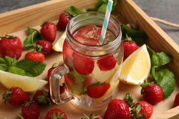 Refreshing drink with strawberries, lemon and mint in mason jar surrounded by fruits in wooden crate on grey table, closeup