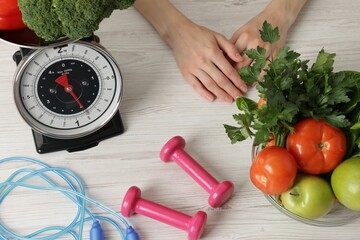 Woman at light wooden table with healthy food, kitchen scale, skipping rope and dumbbells, top view