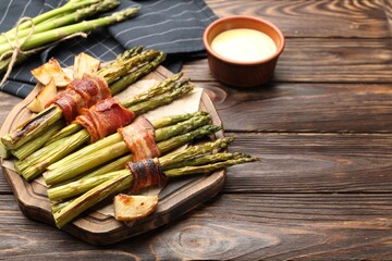 Tasty cooked asparagus served with bacon and sauce on wooden table. Space for text