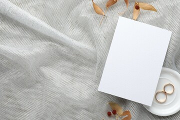Wedding invitation card, golden rings and dry leaves on grey tulle cloth, top view. Space for text