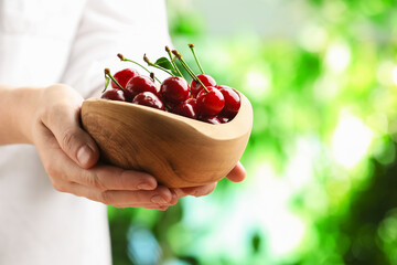 Woman with bowl of fresh cherries against blurred green background, closeup. Space for text