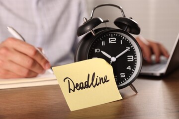 Paper note with word Deadline on alarm clock and man working at wooden table, selective focus