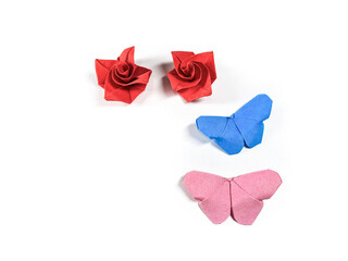 Origami paper butterfly in pink and blue with red rose petals on a white background. Origami butterfly flying towards blooming flowers welcoming spring. Symbolizing hope and happiness for a new life.