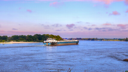 Large cargo ship sailing on the Rhine River under a pastel-colored sky, transporting goods through...
